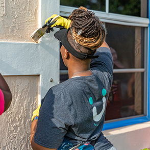 A woman wearing a gray shirt and yellow gloves paints a house.