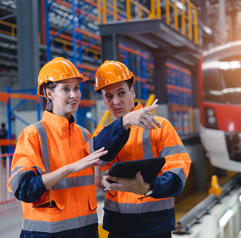 Two engineers in hard hats and safety vests are looking at a tablet in front of a train.